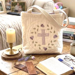 May include: A cream-colored quilted tote bag featuring a central embroidered design of a light purple gingham cross surrounded by a wreath of green leaves and small purple flowers, with two matching fabric handles attached to the top.
