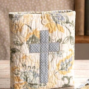 May include: A quilted fabric book cover featuring a cream background with a yellow and blue floral pattern. A gray and white checkered cross is appliqued on the front center. The cover is secured with a small white button and a fabric loop.