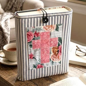 May include: A fabric book cover featuring a blue and white vertical striped pattern. The cover has a black elastic closure with a round button. A rectangular patch is centered on the front, displaying a pink cross over a floral rose print.