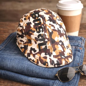 May include: A camouflage print flat cap with the word "Diesel" in black block letters. The cap is resting on a folded pair of blue jeans, with sunglasses and a coffee cup in the background.