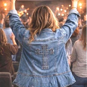 May include: A light blue denim jacket with a cross design on the back. The jacket has a ruffled hem and is worn by a person with their arms raised. The background shows a crowd of people and stage lights.