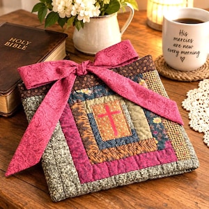 May include: A square quilted fabric book cover featuring a log cabin pattern with various floral and geometric prints in shades of brown, green, and pink. A bright pink paisley fabric ribbon is tied in a bow at the top. A cross is centered.