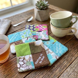 May include: A set of square fabric coasters featuring patchwork designs in shades of green, blue, brown, and pink. The coasters display printed text including "be still and know," "Washed in the Blood," "Giving it to God," and "choose faith."