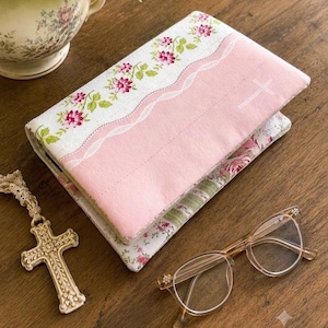May include: A rectangular book cover featuring a pink fabric section with a white cross and a white fabric section with a pink floral pattern, separated by a white wavy trim. A beige cross pendant on a beaded chain and clear glasses are nearby.