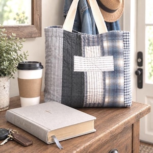 May include: A quilted tote bag featuring vertical panels of blue and gray fabric with a central cross applique. The bag has two cream-colored fabric handles and a patchwork design consisting of stripes, plaid, and gingham patterns in muted tones.