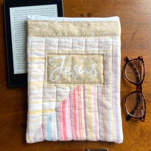 May include: A quilted fabric e-reader case with the word "Jesus" embroidered on the front. The case features a patchwork design with pastel stripes and a cream-colored zipper. A pen, glasses, and an e-reader are also visible.