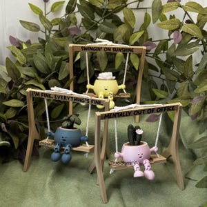 May include: Three whimsical planter figurines on wooden swings. The planters are yellow, blue, and pink, each with a different plant and facial expression. Text on the swings reads "SWINGIN' ON SUNSHINE", "I'M FINE, EVERYTHING'S FINE", and "MENTALLY OUT OF OFFICE".