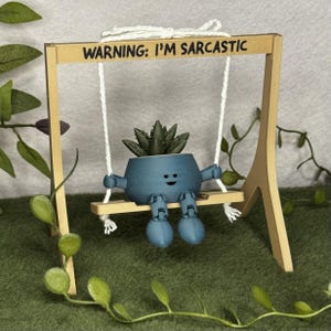 Op de afbeelding: Een blauwe 3D-geprinte plantenpot in de vorm van een figuurtje met armen en benen, met een groene vetplant, zittend op een kleine houten schommel. Het frame is lichtbruin en heeft de zwarte tekst WARNING: I'M SARCASTIC op de bovenste balk.