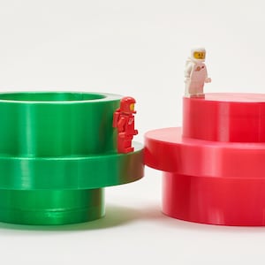 May include: Two large, colorful, plastic toy structures, one green and one red, with a small astronaut figure on each. The green structure is cylindrical with a wide base. The red structure is a tiered, circular design. The astronauts are red and white.