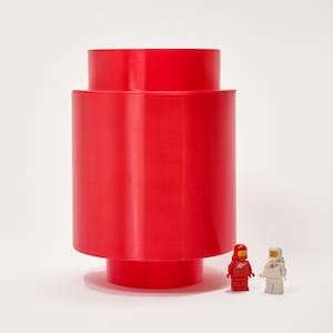 May include: A large, bright red cylindrical object with a stepped design, resembling a building block, stands on a white surface. Two small Lego astronaut figures, one red and one white, are positioned in front of it.