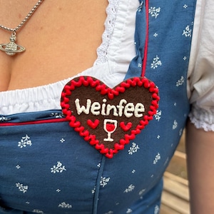 Könnte beinhalten: Eine herzförmige braune Filzbrosche mit rotem Wellenrand. Der Schriftzug "Weinfee" ist in weißem Garn über einem weißen Weinglas-Symbol eingestickt. Unter dem Text befinden sich zwei kleine rote Herzen auf der braunen Filzoberfläche.