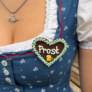 Könnte beinhalten: Eine herzförmige braune Brosche mit hellgrünem Rand, verziert mit dem weißen Schriftzug 'Prost' und einer kleinen gelben Bierkrug-Grafik, befestigt an einem blauen Kleid mit weißem Blumenmuster und einer weißen Bluse mit Spitzenbesatz.