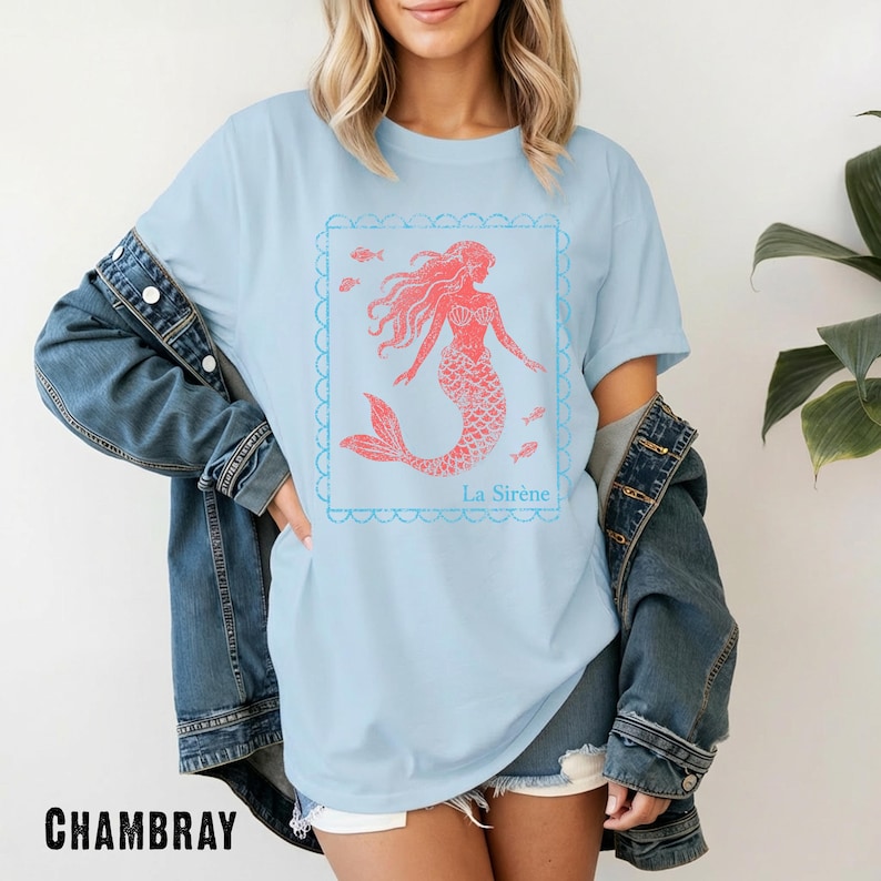 May include: Light blue t-shirt featuring a coral-colored mermaid design within a scalloped frame. The mermaid has flowing hair and a fish tail, with the text "La Sir&egrave;ne" below. The shirt is made of a soft material.