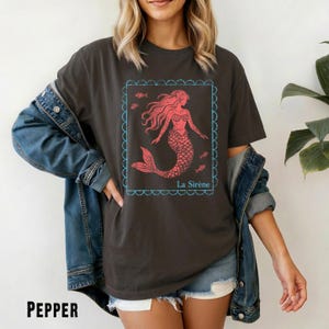 May include: A dark gray t-shirt with a red mermaid graphic inside a blue scalloped frame. The mermaid has flowing hair and a fish tail. The text "La Sir&egrave;ne" is below the graphic. The word "PEPPER" is at the bottom.