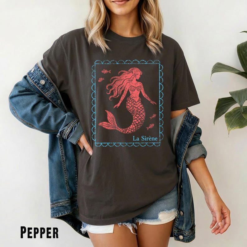 May include: A dark gray t-shirt with a red mermaid graphic inside a blue scalloped frame. The mermaid has flowing hair and a fish tail. The text "La Sir&egrave;ne" is below the graphic. The word "PEPPER" is at the bottom.