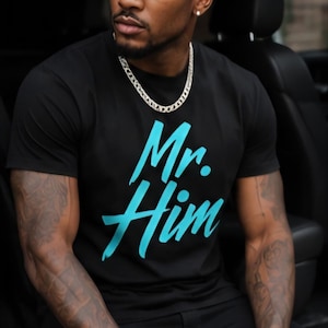 May include: Black t-shirt with the text "Mr. Him" in turquoise. The person is wearing a silver chain and a watch. The person is sitting in a car.
