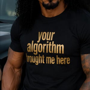 May include: A black short-sleeved t-shirt featuring the gold text "your algorithm brought me here" printed on the chest. The shirt has a crew neckline and a standard fit, showcasing a clear and legible graphic design on the front of the garment.