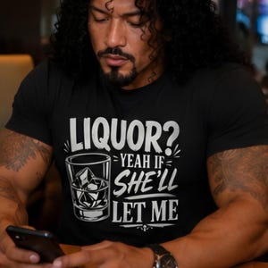 May include: A black short-sleeved t-shirt featuring a white graphic print of a glass filled with ice cubes. The text on the shirt reads: LIQUOR? YEAH IF SHE'LL LET ME. The design is centered on the chest area of the garment.