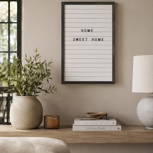 May include: A rectangular wall-mounted letter board with a black frame and white background featuring horizontal grooves. The black plastic letters spell out the phrase HOME SWEET HOME in the center of the board, arranged in two distinct lines.