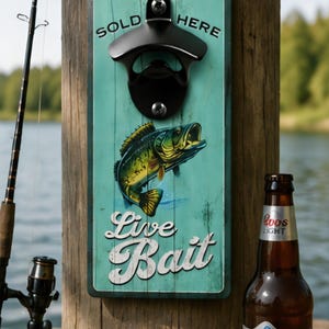 May include: A rectangular wall-mounted bottle opener featuring a teal wood-grain background with a graphic of a green fish. The text reads SOLD HERE and Live Bait. A black metal bottle opener is attached to the top center of the wooden plaque.