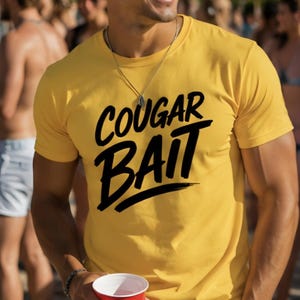 May include: Yellow t-shirt with the text "COUGAR BAIT" in black script. The shirt is worn by a person wearing sunglasses and a necklace, holding a red cup. The background is blurred with other people.