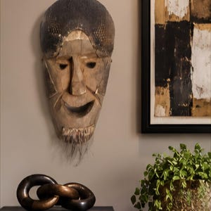 May include: A carved wooden wall mask featuring a dark textured forehead, a light-toned face with a carved beard, and an open mouth. Below, a dark metal chain link sculpture rests on a book titled AFRICA THE ART OF CULTURE.