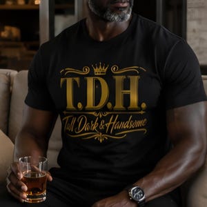 May include: A black short-sleeved t-shirt featuring gold-colored text that reads T.D.H. with a crown icon above it and the words Tall Dark & Handsome written in a cursive script below, all centered on the chest of the garment.