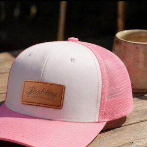 May include: A pink and white trucker hat with a leather patch that reads "Lashley". The hat has a pink brim and mesh back. A beige mug sits next to the hat on a wooden surface.