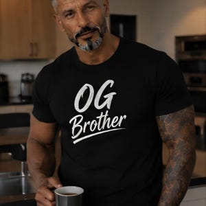 May include: A black short-sleeved t-shirt featuring the white text OG Brother printed in a bold, stylized font across the chest. The garment is made of a solid black fabric and displays the text clearly in the center of the front panel.