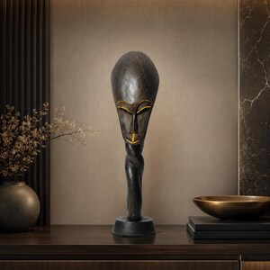 May include: A dark brown wooden sculpture featuring an elongated, oval-shaped head with stylized facial features, including almond-shaped eyes and a narrow nose highlighted with gold paint, resting on a twisted neck and a circular base.