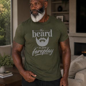 May include: An olive green short-sleeved t-shirt featuring a graphic of a stylized beard and the text "the beard is just foreplay" printed in a light gray color across the chest. The shirt has a crew neckline and a standard fit design.