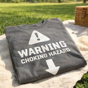 Può includere: Una maglietta grigio scuro con la scritta bianca "WARNING CHOKING HAZARD", un simbolo di punto esclamativo bianco all'interno di un triangolo e una freccia rivolta verso il basso. La maglietta è piegata e appoggiata su una superficie chiara.