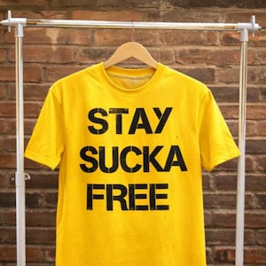 Op de afbeelding: Een felgeel t-shirt met de tekst "STAY SUCKA FREE" in grote, zwarte letters. Het t-shirt hangt aan een houten hanger tegen een achtergrond van een rode bakstenen muur.