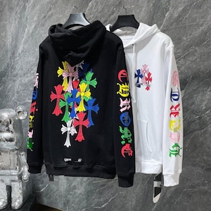 May include: Black and white hoodies with colorful cross designs. The black hoodie features a cluster of crosses on the back and text on the sleeves. The white hoodie has a cross design on the chest and text on the sleeves.