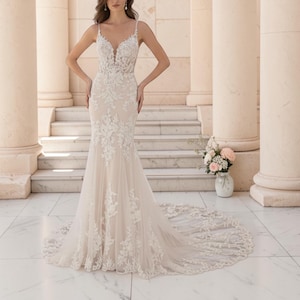 Puede incluir: Un vestido de novia blanco con silueta de sirena. El vestido presenta delicados detalles de encaje en el corpiño y la falda, con tirantes finos y un escote en V. El vestido tiene una larga cola y se muestra en un entorno al aire libre.