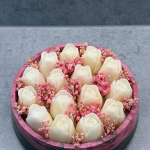May include: A round container with a black and pink marbled exterior filled with multiple white tulip-shaped candles. The spaces between the candles are filled with small, dried pink baby's breath flowers, creating a dense floral arrangement.