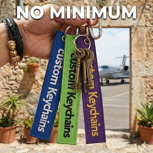 May include: A hand holds a set of keys attached to three custom keychains. The keychains are blue, green, and purple, embroidered with "custom Keychains" in white. The text "NO MINIMUM" is at the top. A bracelet with gold lion heads is visible.