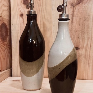 May include: Two ceramic oil and vinegar bottles with stainless steel pour spouts. One bottle is black with a tan and white diagonal stripe, the other is white with a black and tan diagonal stripe. The bottles are on a wooden surface.