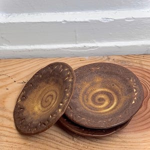 May include: A stack of three round ceramic plates with a dark brown and golden yellow finish. Each plate features a central spiral pattern and a decorative border of small, evenly spaced indentations along the outer rim.