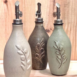 May include: Three ceramic olive oil bottles with stainless steel spouts. The bottles are in shades of beige, brown, and green, each featuring an embossed olive branch design. They are arranged on a wooden surface.