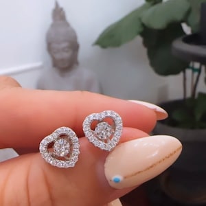 May include: A pair of silver heart-shaped earrings with a clear crystal in the center of each heart. The earrings are surrounded by smaller clear crystals.