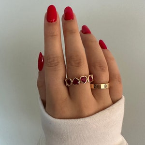 May include: Two gold rings on a hand with red nail polish. The top ring has a row of red heart-shaped stones. The bottom ring has the inscription "Cartier" on it.