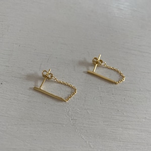 May include: A pair of gold earrings with a small bar and a delicate chain hanging from it.