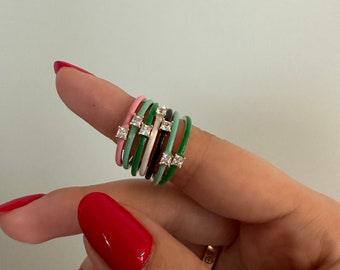May include: A selection of colourful enamel rings, each featuring square-cut gemstones. The rings are in shades of pink, green, black, and white. The rings are stacked on a finger, displaying the variety of colours and designs.