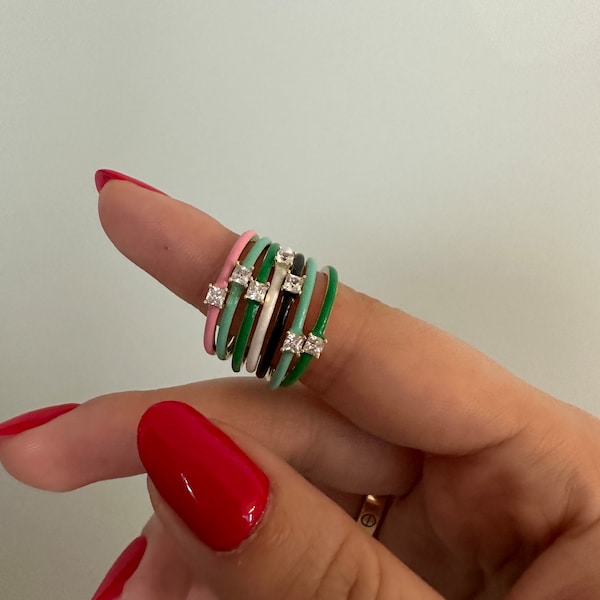 May include: A selection of colourful enamel rings, each featuring square-cut gemstones. The rings are in shades of pink, green, black, and white. The rings are stacked on a finger, displaying the variety of colours and designs.