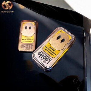 May include: Two rectangular metal Ghost 2025 collectibles with a gold ghost design on a yellow background. The collectibles have the text "HALOO PLAYER X ZETE INFINITY" and are on a reflective black surface.