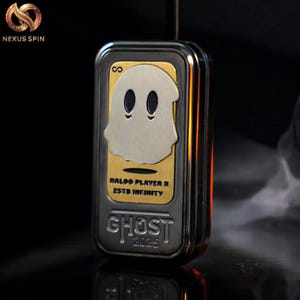 May include: A rectangular, metallic object with a gold and silver finish. The front features a ghost design with the text "HALOO PLAYER X ZSTB INFINITY" and "GHOST 2025". The object has a sleek, modern design.
