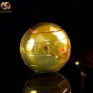 May include: A gold-colored, spherical fidget spinner with horizontal grooves. The spinner reflects the surrounding environment, creating a mirrored effect. The logo "Nexus Spin" is visible in the upper left corner.