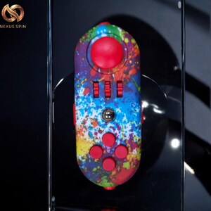 May include: A colorful fidget toy with a vibrant splatter paint design in red, blue, yellow, and green. It features a large red joystick, three red switches, a silver button, and five red buttons. The product is branded with the text "Nexus Spin".