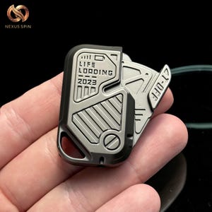 May include: A metallic gray and black pocket tool with the text "LIFE LOADING 2023" and "21-OFF". The tool has a futuristic, industrial design with a key ring hole and a hinged blade.
